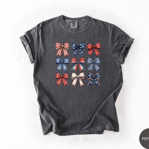 May include: A charcoal gray short-sleeve t-shirt featuring a grid of nine decorative bows in various red, white, and blue patterns, including stars, stripes, and solid colors. The shirt has a crew neckline and a relaxed, casual fit.