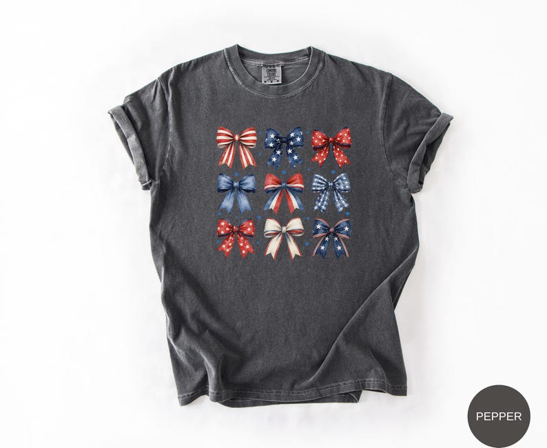 May include: A charcoal gray short-sleeve t-shirt featuring a grid of nine decorative bows in various red, white, and blue patterns, including stars, stripes, and solid colors. The shirt has a crew neckline and a relaxed, casual fit.