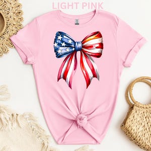 May include: Light pink t-shirt featuring a watercolor bow design in red, white, and blue, resembling the American flag. The bow has a blue center with white stars and red and white striped ribbons. The shirt is knotted at the waist.