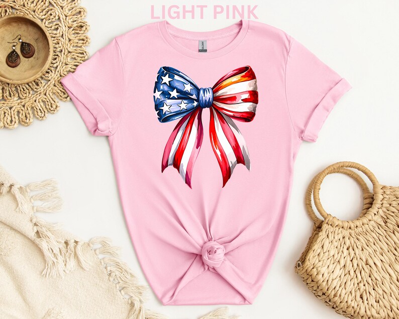 May include: Light pink t-shirt featuring a watercolor bow design in red, white, and blue, resembling the American flag. The bow has a blue center with white stars and red and white striped ribbons. The shirt is knotted at the waist.