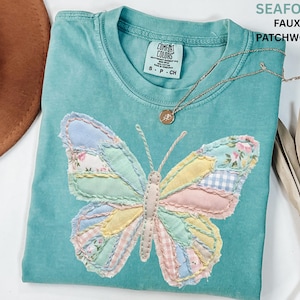 May include: Seafoam green t-shirt with a faux patchwork butterfly design. The butterfly features various pastel fabrics, including floral and gingham patterns. The shirt has a crew neck and short sleeves. A gold necklace with a pendant is also visible.