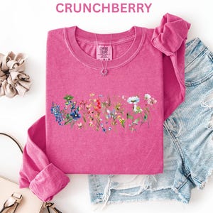 May include: A pink long-sleeve shirt with a floral design, the word "CRUNCHBERRY" is at the top. The shirt is paired with denim shorts, a scrunchie, and a purse. A silver necklace with a sun pendant is visible.