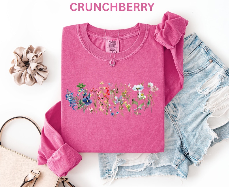 May include: A pink long-sleeve shirt with a floral design, the word "CRUNCHBERRY" is at the top. The shirt is paired with denim shorts, a scrunchie, and a purse. A silver necklace with a sun pendant is visible.