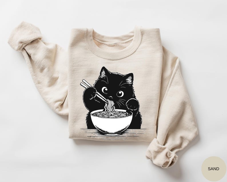 Black cat illustration sweatshirt featuring a cute cat eating noodles from a bowl on a sand color crewneck sweatshirt
