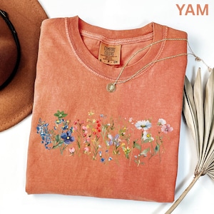 May include: A coral-colored t-shirt with a crew neck and a floral design across the front. The design features a variety of colorful wildflowers. A gold necklace with a pendant is draped over the shirt. The tag reads "Comfort Colors".