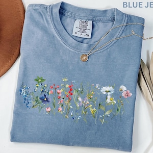 May include: A blue jean colored t-shirt with a floral design across the front. The shirt has a crew neck and short sleeves. A gold necklace with a pendant is draped across the shirt. The shirt has the Comfort Colors brand tag.