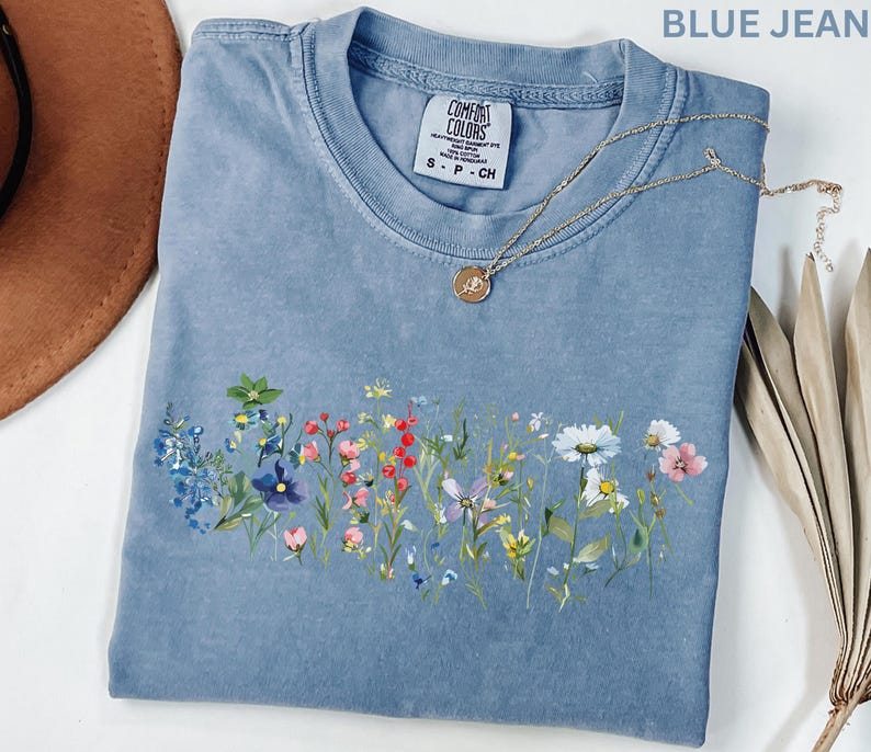 May include: A blue jean colored t-shirt with a floral design across the front. The shirt has a crew neck and short sleeves. A gold necklace with a pendant is draped across the shirt. The shirt has the Comfort Colors brand tag.