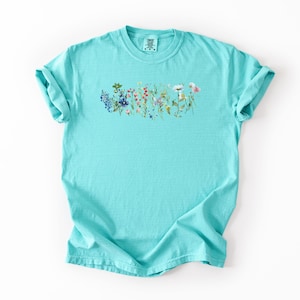 May include: A turquoise t-shirt featuring a floral design across the chest. The design includes a variety of colorful wildflowers in shades of blue, red, pink, and yellow. The shirt has short sleeves and a crew neck.