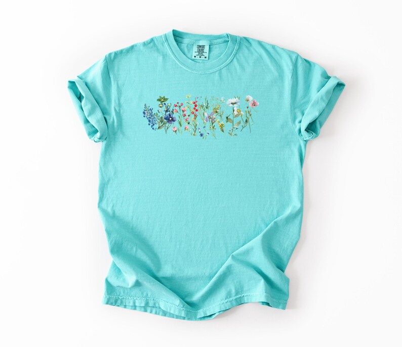 May include: A turquoise t-shirt featuring a floral design across the chest. The design includes a variety of colorful wildflowers in shades of blue, red, pink, and yellow. The shirt has short sleeves and a crew neck.