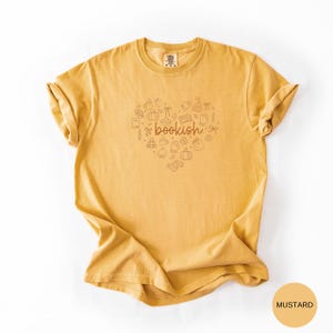 May include: A mustard yellow short-sleeve t-shirt featuring a central graphic of a heart shape composed of small line-art icons including books, pumpkins, and mugs. The word "bookish" is printed in a cursive brown font in the center.