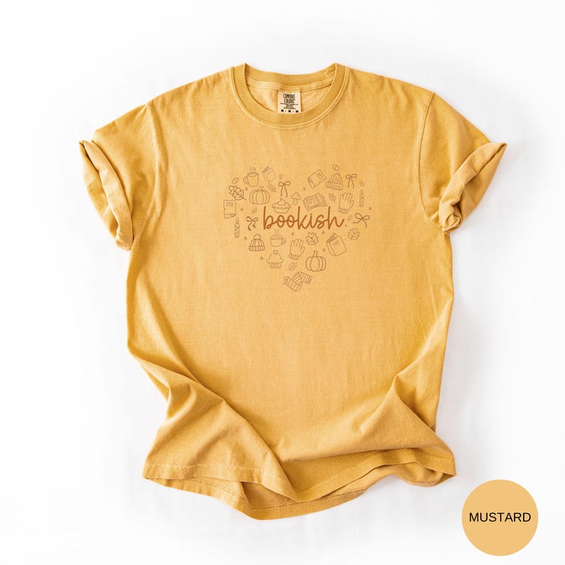May include: A mustard yellow short-sleeve t-shirt featuring a central graphic of a heart shape composed of small line-art icons including books, pumpkins, and mugs. The word "bookish" is printed in a cursive brown font in the center.