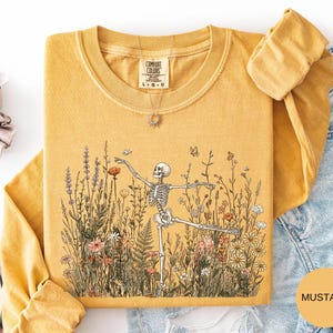 Puede incluir: Una camiseta de manga larga color amarillo mostaza con un estampado gr&aacute;fico de un esqueleto bailando en medio de un campo de flores silvestres y mariposas. La camiseta tiene cuello redondo y una etiqueta de la marca Comfort Colors.