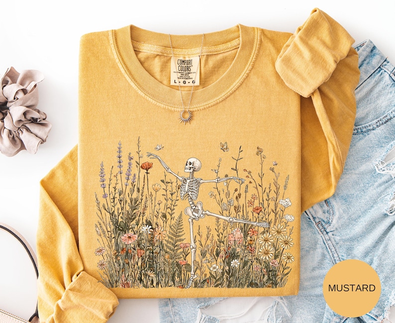 Puede incluir: Una camiseta de manga larga color amarillo mostaza con un estampado gr&aacute;fico de un esqueleto bailando en medio de un campo de flores silvestres y mariposas. La camiseta tiene cuello redondo y una etiqueta de la marca Comfort Colors.