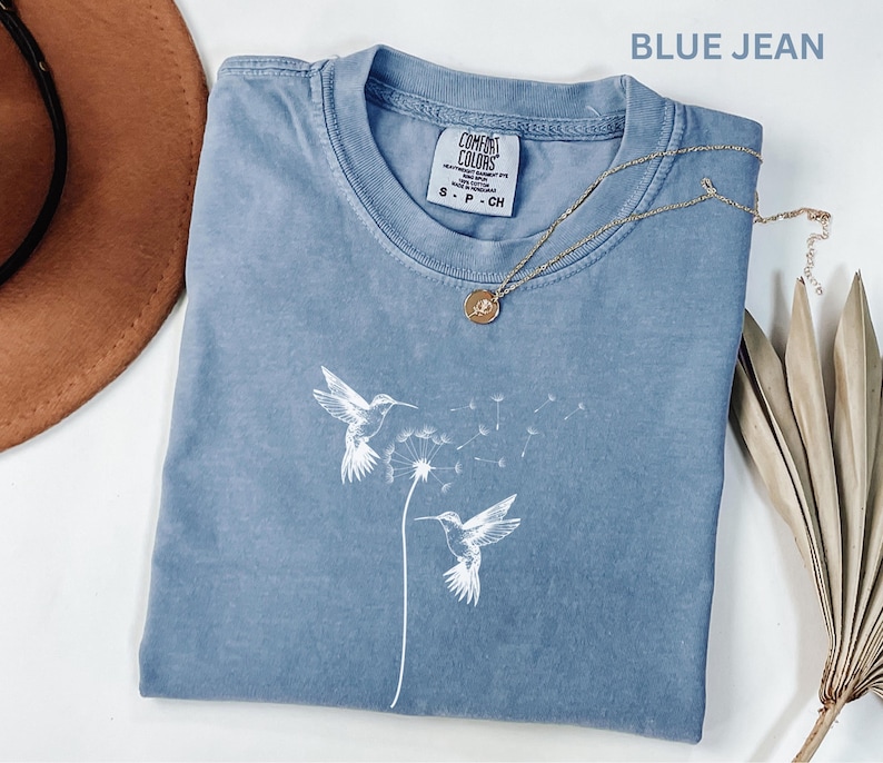 a blue t-shirt with a white bird design, a brown hat, a necklace, and some dried leaves, all arranged on a white surface.