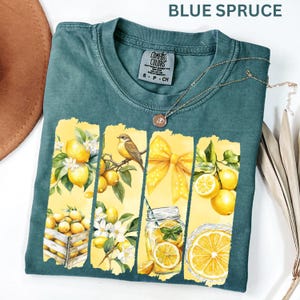 May include: Blue Spruce Comfort Colors t-shirt with a lemon-themed graphic. The graphic features lemons, a bird, a yellow bow, and a jar of lemonade. A gold necklace is draped across the shirt. The shirt is size S-P-CH.