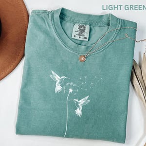 a light green t-shirt with a white bird design, a necklace, a brown hat, and some dried leaves on a white background.