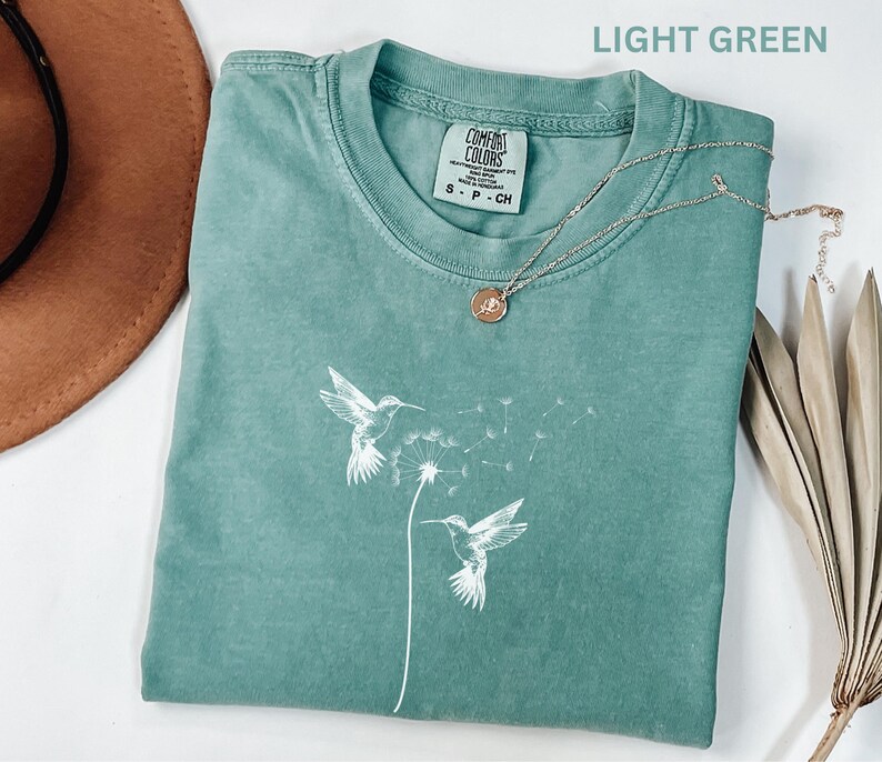 a light green t-shirt with a white bird design, a necklace, a brown hat, and some dried leaves on a white background.