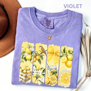 May include: Violet Comfort Colors t-shirt with a lemon-themed design. The graphic includes lemons, a bird, a bow, and a jar of lemonade. A gold necklace and a brown hat are also visible.