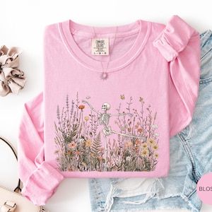 Puede incluir: Una sudadera de cuello redondo y manga larga en color rosa claro con un estampado gr&aacute;fico de un esqueleto bailando entre flores silvestres y mariposas. La prenda incluye una etiqueta de la marca Comfort Colors en el cuello, talla L-G-G.