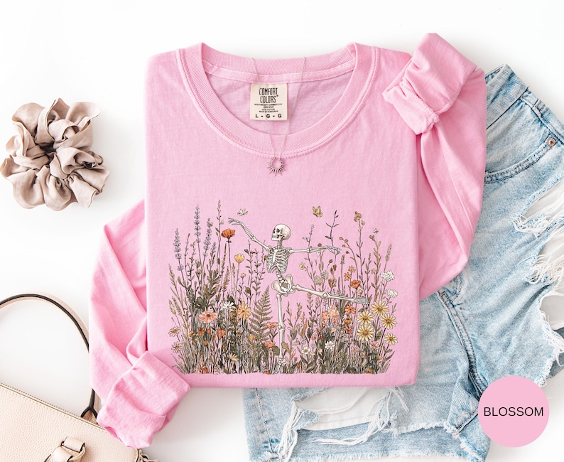 Puede incluir: Una sudadera de cuello redondo y manga larga en color rosa claro con un estampado gr&aacute;fico de un esqueleto bailando entre flores silvestres y mariposas. La prenda incluye una etiqueta de la marca Comfort Colors en el cuello, talla L-G-G.