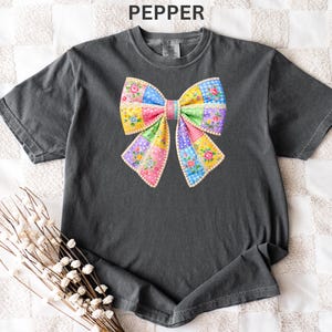 May include: A charcoal grey t-shirt with a vibrant bow graphic. The bow is constructed from patchwork fabric, incorporating floral and polka dot patterns in pink, yellow, blue, and green. The word "PEPPER" is printed at the top.