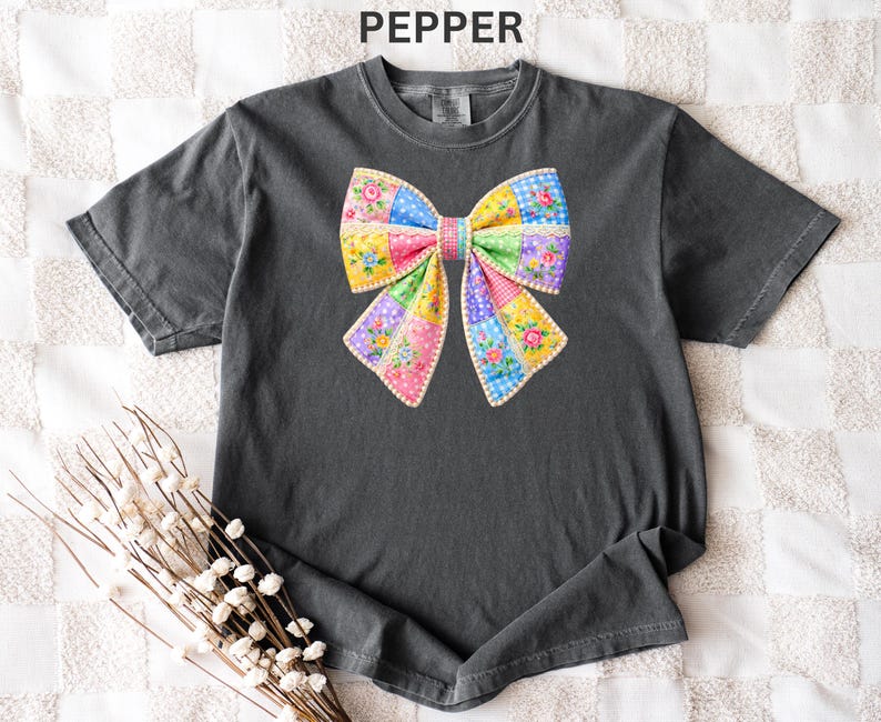 May include: A charcoal grey t-shirt with a vibrant bow graphic. The bow is constructed from patchwork fabric, incorporating floral and polka dot patterns in pink, yellow, blue, and green. The word "PEPPER" is printed at the top.
