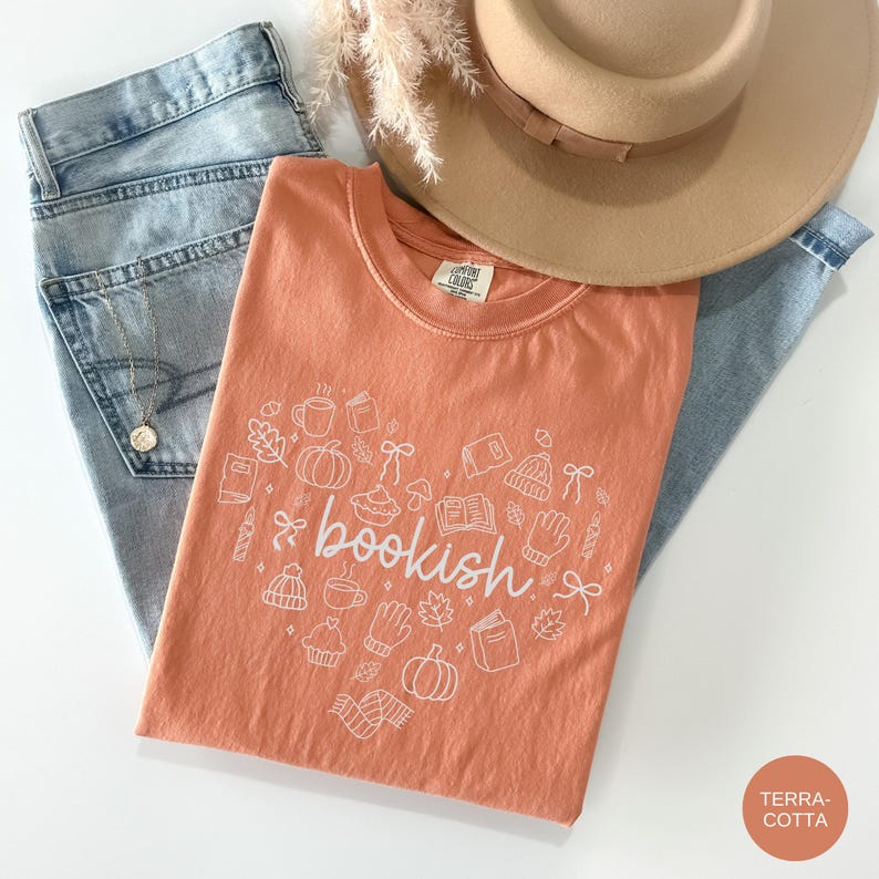 May include: A terracotta t-shirt featuring a white graphic print of the word bookish surrounded by small line drawings of pumpkins, books, mugs, leaves, and winter accessories, paired with light blue denim jeans, a tan hat, and a gold necklace.