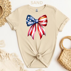 May include: A sand-colored t-shirt featuring a watercolor bow design. The bow is a patriotic mix of red and white stripes with blue and white stars, resembling the American flag. The shirt is knotted at the waist.