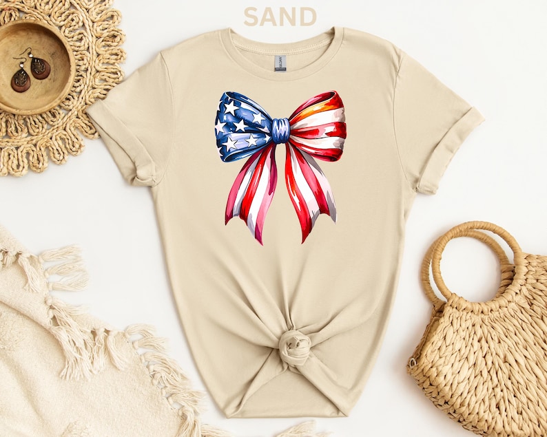 May include: A sand-colored t-shirt featuring a watercolor bow design. The bow is a patriotic mix of red and white stripes with blue and white stars, resembling the American flag. The shirt is knotted at the waist.