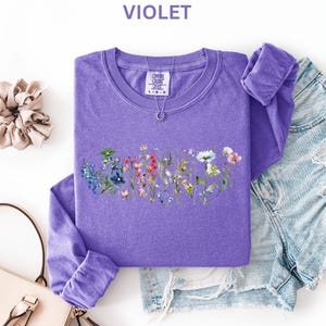 May include: A violet long-sleeve shirt with a floral design, a scrunchie, a purse, and denim shorts. The shirt has a crew neck and the word "VIOLET" at the top. A necklace with a sun pendant is on the shirt.