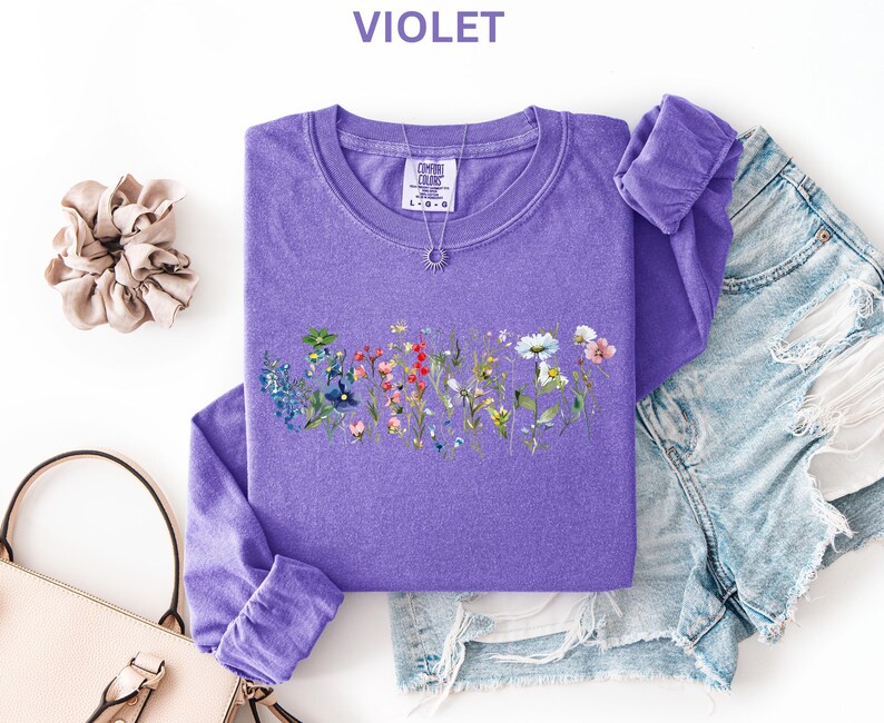 May include: A violet long-sleeve shirt with a floral design, a scrunchie, a purse, and denim shorts. The shirt has a crew neck and the word "VIOLET" at the top. A necklace with a sun pendant is on the shirt.