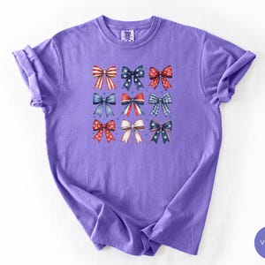 May include: A violet short-sleeved t-shirt featuring a central graphic print of nine decorative bows arranged in a three-by-three grid. The bows display various red, white, and blue patterns, including stripes, polka dots, stars, and plaid designs.