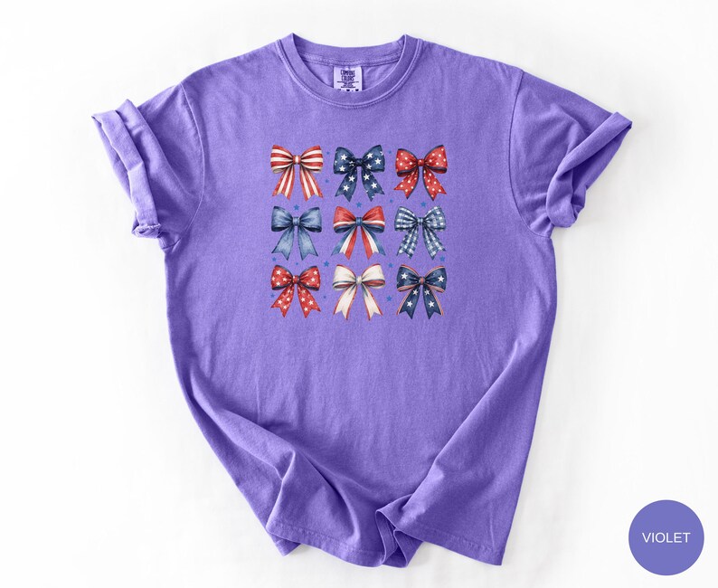 May include: A violet short-sleeved t-shirt featuring a central graphic print of nine decorative bows arranged in a three-by-three grid. The bows display various red, white, and blue patterns, including stripes, polka dots, stars, and plaid designs.