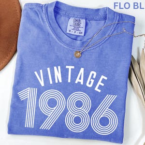 May include: A blue t-shirt with the text "VINTAGE 1986" in white. The shirt has a crew neck and short sleeves. A gold necklace with a pendant is draped across the shirt. The shirt is folded on a white surface.