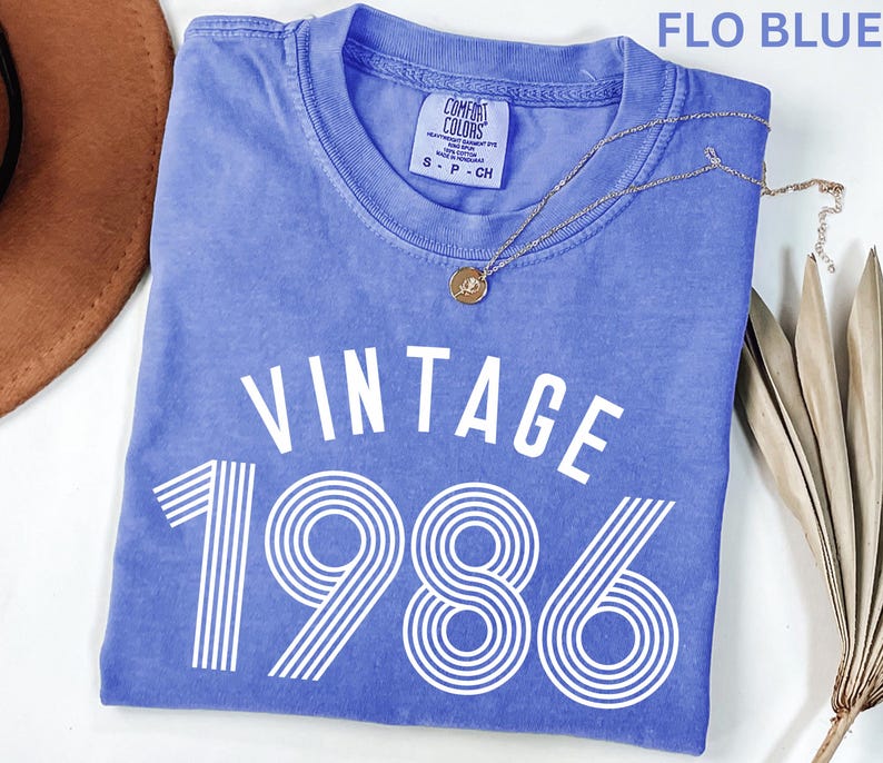 May include: A blue t-shirt with the text "VINTAGE 1986" in white. The shirt has a crew neck and short sleeves. A gold necklace with a pendant is draped across the shirt. The shirt is folded on a white surface.