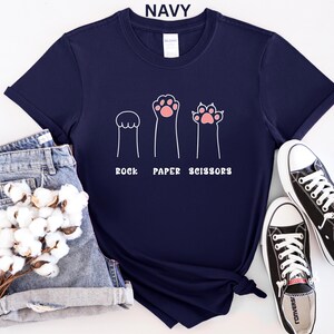 Puede incluir: Camiseta azul marino con un dise&ntilde;o l&uacute;dico de "Piedra, Papel o Tijeras" con ilustraciones de patas. El dise&ntilde;o incluye tres figuras en forma de pata en blanco con acentos rosas. La camiseta se combina con pantalones cortos vaqueros y zapatillas negras.