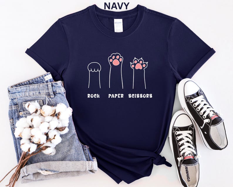 Puede incluir: Camiseta azul marino con un dise&ntilde;o l&uacute;dico de "Piedra, Papel o Tijeras" con ilustraciones de patas. El dise&ntilde;o incluye tres figuras en forma de pata en blanco con acentos rosas. La camiseta se combina con pantalones cortos vaqueros y zapatillas negras.