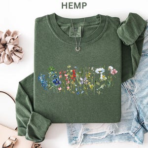 May include: A green long-sleeve shirt with a floral design across the chest. The design features a variety of colorful wildflowers. A silver necklace with a sun pendant is visible. The word "HEMP" is printed at the top.