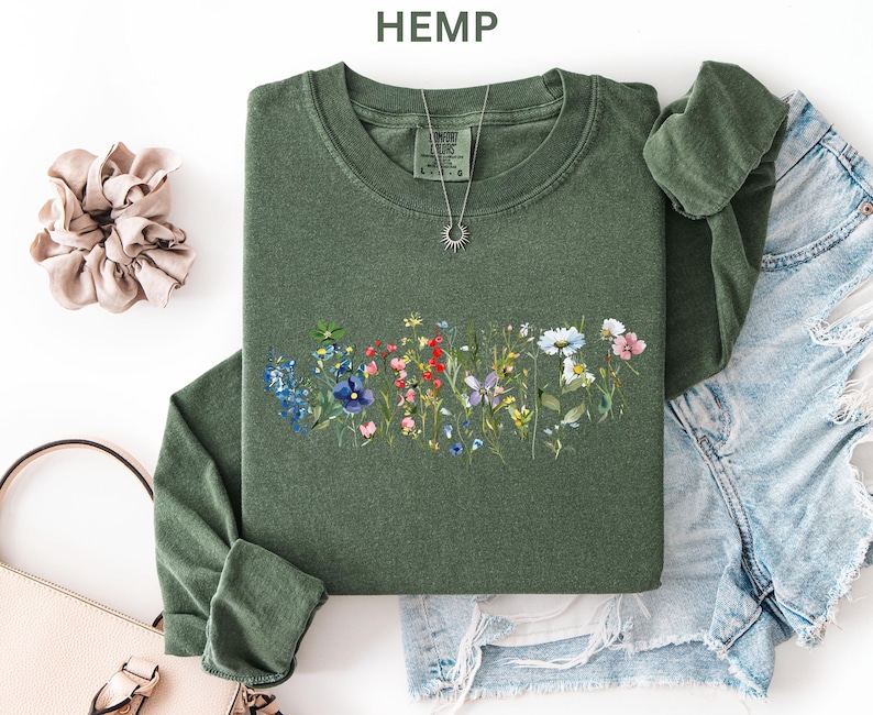 May include: A green long-sleeve shirt with a floral design across the chest. The design features a variety of colorful wildflowers. A silver necklace with a sun pendant is visible. The word "HEMP" is printed at the top.