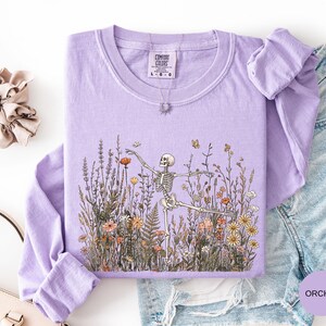 Puede incluir: Una camiseta de manga larga color morado orqu&iacute;dea con un estampado gr&aacute;fico de un esqueleto bailando en medio de un campo de flores silvestres. La camiseta tiene cuello redondo y una etiqueta de la marca Comfort Colors en el cuello.