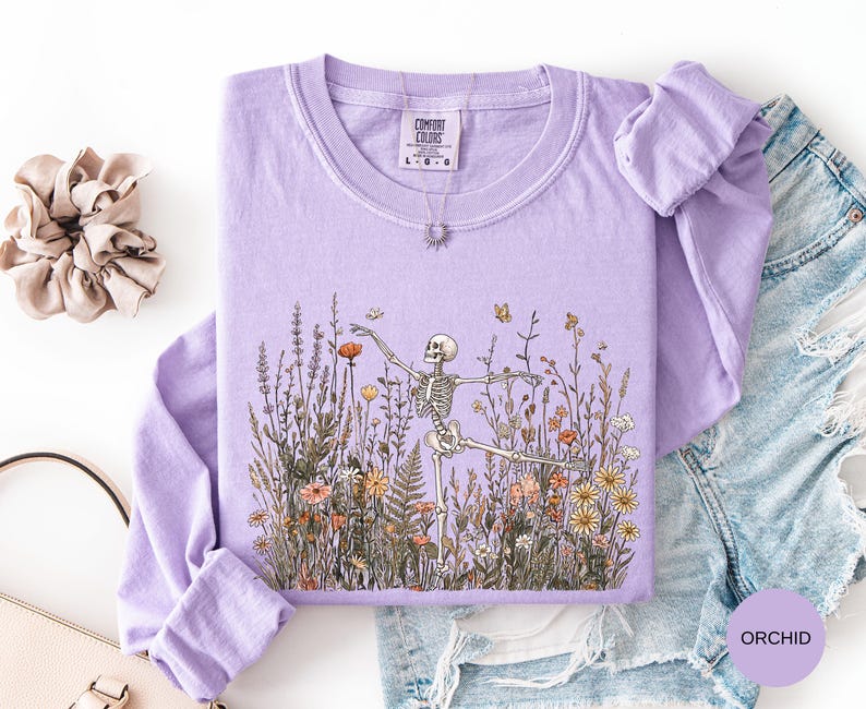Puede incluir: Una camiseta de manga larga color morado orqu&iacute;dea con un estampado gr&aacute;fico de un esqueleto bailando en medio de un campo de flores silvestres. La camiseta tiene cuello redondo y una etiqueta de la marca Comfort Colors en el cuello.