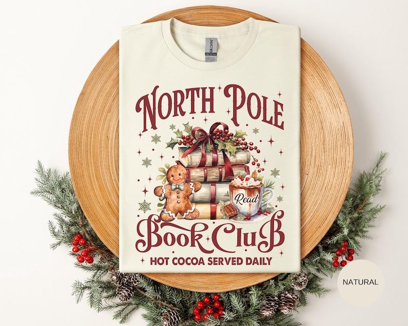 May include: A cream-colored t-shirt featuring a graphic with the text North Pole Book Club and Hot Cocoa Served Daily. The design includes a stack of books tied with a red ribbon, a gingerbread person, and a mug of hot cocoa labeled Read.