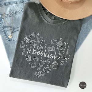 May include: A charcoal gray crew neck t-shirt featuring a white line art graphic in the shape of a heart. The design includes the word bookish in the center surrounded by small illustrations of pumpkins, books, mugs, leaves, mittens, and hats.