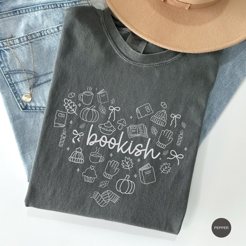May include: A charcoal gray crew neck t-shirt featuring a white line art graphic in the shape of a heart. The design includes the word bookish in the center surrounded by small illustrations of pumpkins, books, mugs, leaves, mittens, and hats.