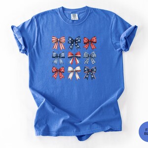 May include: A short-sleeved, crew-neck t-shirt in a vibrant blue color. The center of the shirt features a grid of nine decorative bows in various patterns, including red and white stripes, blue with white stars, and red with white polka dots.