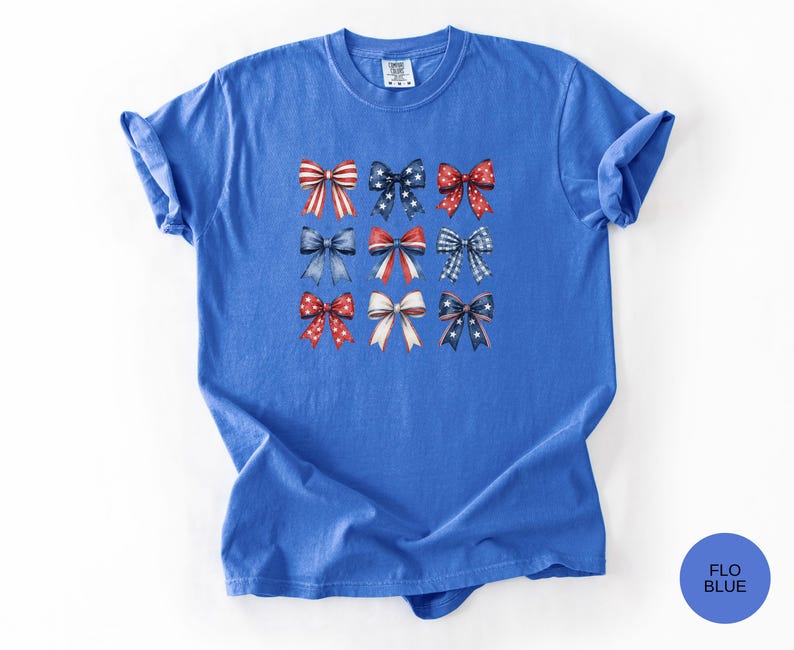 May include: A short-sleeved, crew-neck t-shirt in a vibrant blue color. The center of the shirt features a grid of nine decorative bows in various patterns, including red and white stripes, blue with white stars, and red with white polka dots.