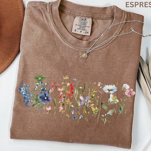 May include: Espresso-colored t-shirt with a floral design across the front. The shirt has a crew neck and short sleeves. A silver necklace with a small pendant rests on the shirt. The shirt has a Comfort Colors tag.