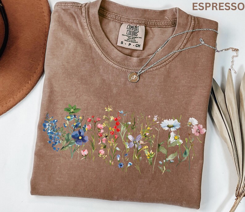 May include: Espresso-colored t-shirt with a floral design across the front. The shirt has a crew neck and short sleeves. A silver necklace with a small pendant rests on the shirt. The shirt has a Comfort Colors tag.