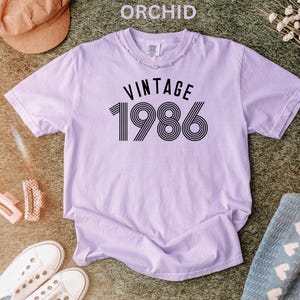 May include: Orchid-colored t-shirt with the text "VINTAGE 1986" in black. The shirt has a distressed neckline and short sleeves. The image also includes a brown hat, white sneakers, and other accessories.