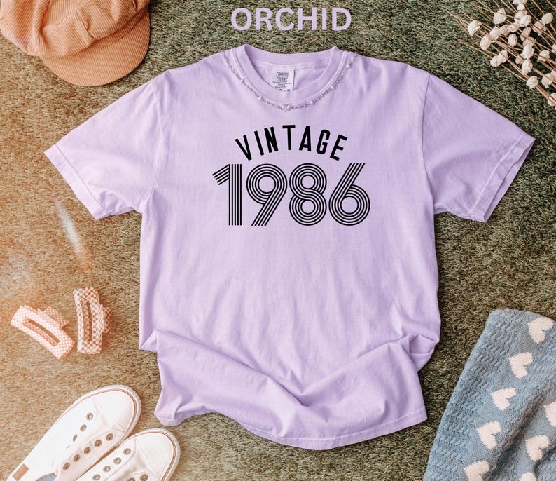 May include: Orchid-colored t-shirt with the text "VINTAGE 1986" in black. The shirt has a distressed neckline and short sleeves. The image also includes a brown hat, white sneakers, and other accessories.
